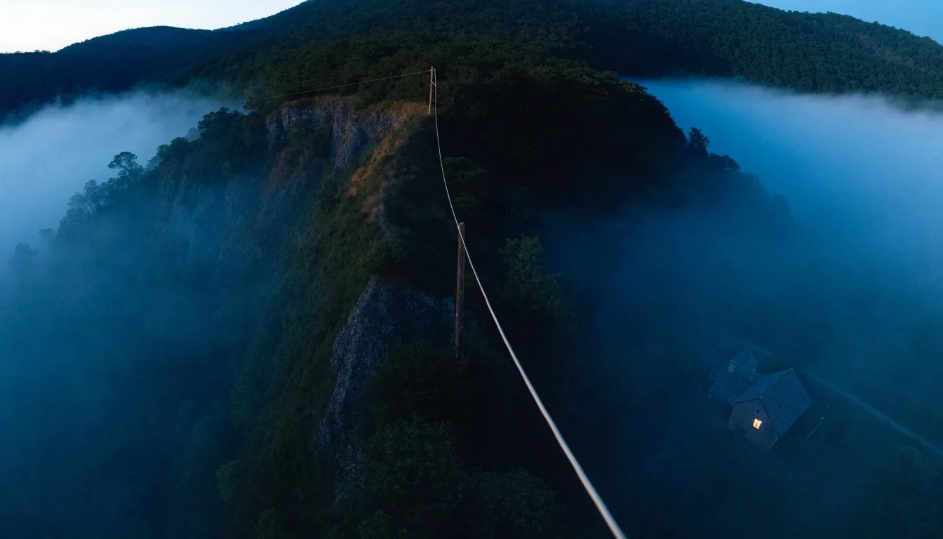 Aerial fibre trunk at dusk above a valley with one lit house and fog behind