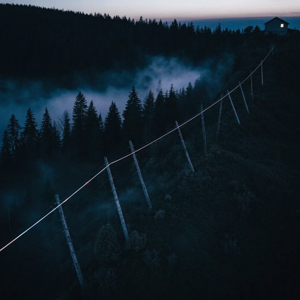 Aerial cable trunk crossing a fogged hillside at dusk with one lit house on the ridge